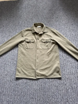 Grön overshirt från Selected Homme - Stilren grön overshirt från Selected Homme i storlek L. Skjortjackan har två bröstfickor med lock och knappar, klassisk krage och tryckknappar framtill. Tillverkad i ett mjukt, kraftigare bomullstyg som passar perfekt för lager på lager.