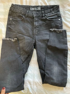 Svarta slitna jeans med fransiga knän - Säljer ett par svarta jeans med fransiga hål vid knäna och klassisk femficksmodell. Jeansen har en smal passform och är tillverkade i stretchigt denimtyg. Perfekta för en avslappnad och trendig look.