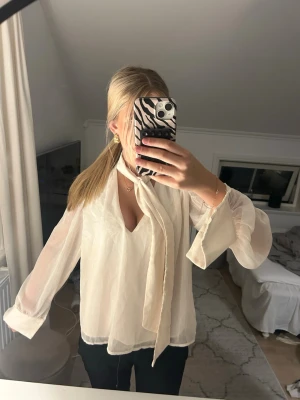 Vit blus med knytband - Stilren vit blus i transparent material med långa ärmar och ett snyggt knytband vid halsen. Blusen har en lös passform och lätt v-ringad urringning. 