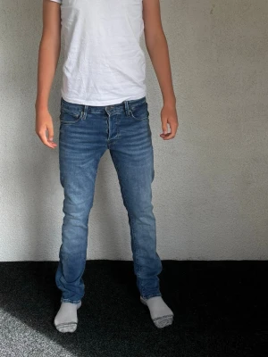 Blå slim fit jeans herr - Säljer ett par blå slim fit jeans med klassisk femficksdesign och normalhög midja. Jeansen har en snygg tvättad look och raka ben. Materialet är denim i bomull med lite stretch för skön passform. Perfekta till en avslappnad stil.