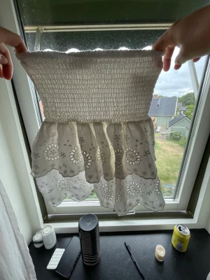 Vit kortkjol med broderade volanger - Söt vit kortkjol med bred smock i midjan och dubbla volanger. Kjolen har broderade blommönster och vågig kant nertill för en romantisk look. Perfekt för varma dagar och enkel att matcha med olika toppar.