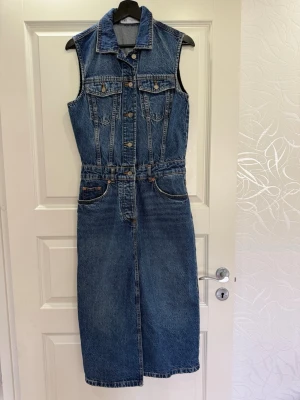 Ärmlös jeansklänning med knappar -  Jeansklänning i blå med krage och knappar framtill. Klänningen har två bröstfickor med lock, framfickor och markerad midja.