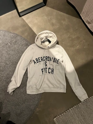 Vit hoodie från Abercrombie & Fitch - Snygg vit hoodie från Abercrombie & Fitch med stor logga i svart text på bröstet. Hoodien har huva med dragsko och en klassisk magficka. Perfekt för en avslappnad och trendig look. Mjuk bomullsblandning och långa ärmar.