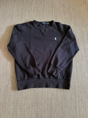 Mörkblå stickad tröja Polo Ralph Lauren - Mörkblå stickad tröja från Polo Ralph Lauren med klassisk rund halsringning och ribbade muddar. Tröjan har den ikoniska broderade loggan i ljusblått på bröstet. Perfekt för en stilren och avslappnad look.