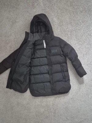 Helly Hansen jacka - Svart pufferjacka från Helly Hansen med huva och dragkedja. Jackan har quiltad design, två sidofickor och HH-logga på bröstet. Tillverkad i syntetmaterial och perfekt för kalla dagar.
