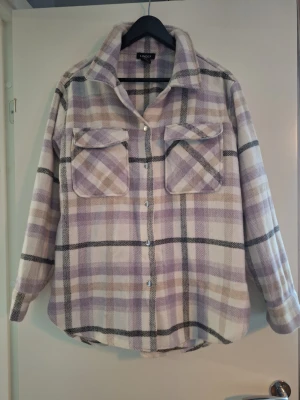 Lila och vit rutig overshirt jacka från Lindex  - Overshirt jacka i mjukt, borstat tyg med rutigt mönster i lila, vitt och beige. Två stora bröstfickor med lock och knappar framtill. Avslappnad passform och klassisk krage. Perfekt lager på lager-plagg för en trendig look. Nypris 599 kr.