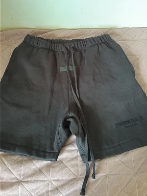  Essentials shorts - Säljer ett par svarta shorts från Essentials med elastisk midja och långa snören framtill. Shortsen har en relaxed fit och diskret logga på vänster ben. Perfekta för chill dagar eller när du vill ha det bekvämt.
