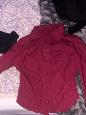 Mörkröd skjorta med knappar - Säljer en mörkröd skjorta med klassisk krage och knappar framtill. Skjortan har lång ärm och är i ett mjukt tyg, perfekt för dig som vill ha en stilren och enkel look. Passar till många olika outfits och är lätt att matcha.
