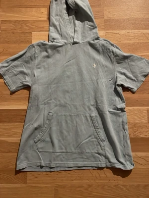 Ljusblå polo hoodie med kort ärm - Snygg ljusblå hoodie från Polo Ralph Lauren med korta ärmar och klassisk känguruficka framtill. Ikoniska broderade loggan på bröstet. Perfekt för en avslappnad och cool stil. Tillverkad i mjukt bomullsmaterial och har en stor huva.
