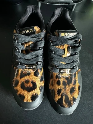 Leopardmönstrade sneakers från BORG - Snygga sneakers från BORG med leopardmönstrad ovandel i syntetmaterial och svarta detaljer. Skorna har svarta snören och en rejäl svart sula med bra grepp. Perfekta för dig som vill sticka ut med ett djurmönster i din outfit.