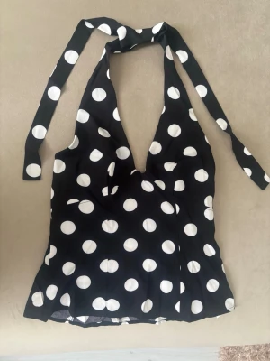 Svart prickig halterneck topp - Snygg svart halterneck topp med stora vita prickar och knyt i nacken. Toppen har djup urringning och är ärmlös, perfekt för en trendig och lekfull stil. Materialet känns lätt och svalt, troligen polyester eller bomull.