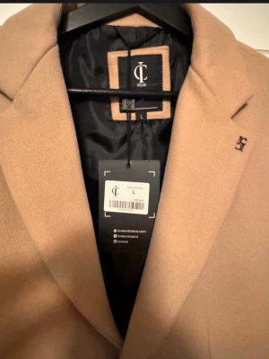 Kamelbrun rock från IC Wear - Stilren kamelbrun rock från IC Wear i storlek L. Klassisk design med breda slag, svart innerfoder och diskret logga på bröstet. Perfekt för dig som vill ha en tidlös och snygg look. Materialet känns mjukt och exklusivt.