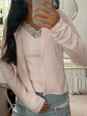 Ljusrosa ribbad kofta med spetsdetalj - Söt ljusrosa kofta i ribbat material med knappar framtill. Under koftan syns ett matchande linne med vit spetskant vid urringningen som ger en romantisk vibe. Perfekt att styla med jeans eller kjol för en chill look.