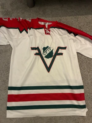 Frölunda Indians hockeytröja S - Säljer en vit och röd hockeytröja från Frölunda Indians med gröna detaljer och klubbens logga på bröstet. Tröjan har snörning i halsen och breda ränder längst ner. Perfekt för dig som vill stötta laget på match!