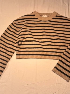 Beige och svart randig cropped tröja - Beige stickad cropped tröja med svarta horisontella ränder. Tröjan har rund halsringning och långa ärmar. Materialet känns mjukt och bekvämt med 6% ull. Lite nopprig därav pris 