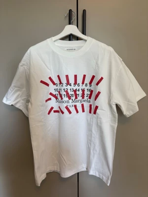 Vit Maison Margiela t-shirt med tryck - Vit t-shirt från Maison Margiela med ikoniskt siffertryck och röda grafiska detaljer på bröstet. Klassisk passform och rund halsringning. Tillverkad i mjuk bomull för en skön känsla och stilren look.