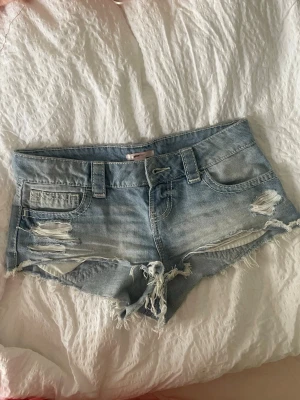 Ljusblå slitna jeansshorts - Säljer ett par lågmidjade ljusblå jeansshorts med slitna detaljer och fransig kant. Shortsen har klassisk femficksmodell, bälteshällor och låg midja.  Perfekta för varma dagar och en avslappnad stil. Men även till sen sommaren o våren stylat med boots 