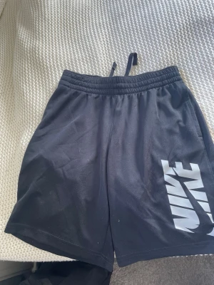 Svarta Nike shorts med logga - Säljer ett par svarta shorts från Nike med stor vit logga på ena benet. Shortsen har elastisk midja med snörning och är tillverkade i ett mjukt och lätt material, perfekta för träning eller chill. Klassisk och sportig look.