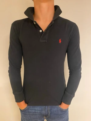 Svart långärmad henley från Polo Ralph Lauren - Säljer en svart långärmad henley från Polo Ralph Lauren med röd broderad logga på bröstet. Slim fit-modell med knappar vid halsen och klassisk krage. Perfekt till jeans för en clean och stilren look.