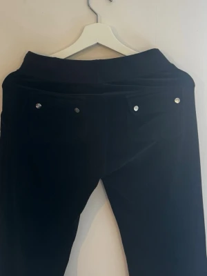 Svarta mjukisbyxor med fickor - Svarta lågmidjade mjukisbyxor i mjukt material från Juicy Couture med resår i midjan och dragsko framtill. Byxorna har raka ben och två bakfickor med silverfärgade knappar. Perfekta för en avslappnad stil. Pris går att diskutera💕💕