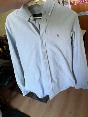 Ljusblå skjorta från Ralph Lauren - Klassisk ljusblå skjorta från Ralph Lauren med button-down krage och broderad logga på bröstet. Skjortan har långa ärmar, vita knappar och är tillverkad i mjuk bomull. Perfekt för en clean och stilren look.