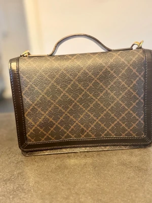 By Malene Birger, Brun handväska  - Snygg handväska i mörkbrunt konstläder med rutmönster i ljusare brunt. Väskan har ett topphandtag, guldfärgade detaljer och en avlång, boxig form. Perfekt för dig som gillar klassisk och stilren design. Den har en liten plats där det är lite sakavt som man kan se på sista bilden.