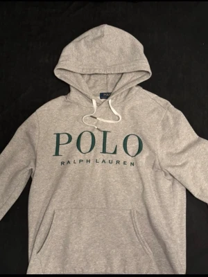 Polo Ralph Lauren Hoodie  - Polo Ralph Lauren hoodie i gråmelerad färg med stort logotryck framtill. Mycket bra skick, sparsamt använd och utan fläckar eller hål. Mjuk och skön kvalitet med justerbar huva och känguruficka. Perfekt till vardags eller en stilren streetwear-look. AUTENTISK!!! 