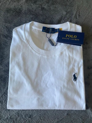 Ralph Lauren T-shirt  - Klassisk vit t-shirt från Polo Ralph Lauren med rund hals och korta ärmar. T-shirten har den ikoniska broderade loggan i mörkblått på bröstet. Tillverkad i mjuk bomull för en skön och stilren look. Inga skador eller hål