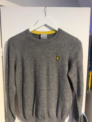 Grå stickad tröja från Lyle & Scott - Står L men passar S. Snygg grå stickad tröja från Lyle & Scott med rund halsringning och gul broderad logga på bröstet. Tröjan har ribbade muddar vid ärmslut och nederkant samt en gul detalj på insidan av kragen. Perfekt för lager på lager.