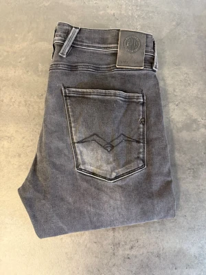 Grå Replay Anbass Jeans, stl 32/34 - Säljer ett par grå replay jeans med tvättad look och klassiska fem fickor. Jeansen har en snygg bakficka med dekorativ söm och normal midja. Perfekta för en avslappnad och trendig stil.