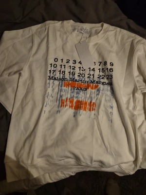 Maison Margiela vit sweatshirt M - Säljer en vit sweatshirt från Maison Martin Margiela i storlek M. Tröjan har tryck med siffror och text på bröstet samt orange och blå detaljer. Rund halsringning och ribbade muddar. Perfekt för dig som gillar streetwear och unika prints.