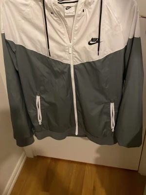 Nike vindjacka vit och grå - Nike vindjacka i vitt och grått med dragkedja framtill och två fickor med dragkedja. Jackan har huva med svarta snören och meshfoder på insidan. Tillverkad i lätt syntetmaterial, perfekt för blåsiga dagar.