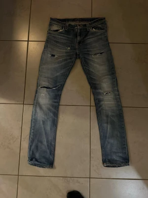 Blå slitna  jeans med hål - Säljer ett par blå skinny jeans med slitningar och hål på både fram- och baksida. Jeansen har en tvättad look, fem fickor och klassisk knappgylf. Perfekta för dig som gillar en avslappnad och trendig stil.