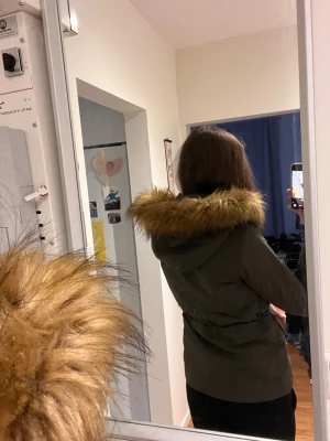 Olivgrön parkas med fuskpäls - Snygg olivgrön parkas med stor luva kantad av brun fuskpäls. Jackan har dragsko i midjan, två stora fickor framtill och stängs med dragkedja och knappar. Perfekt för kalla dagar och ger en trendig look.