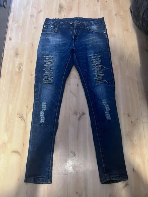 Blå slitna skinny jeans med hål - Säljer ett par blå skinny jeans med slitningar och hål på både fram- och baksida. Jeansen har klassisk femficksmodell och är tillverkade i denim med stretch. Perfekta för en avslappnad och trendig look.