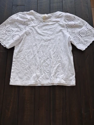 Vit blus med broderade ärmar H&M - Superfin vit blus från H&M med korta, vida ärmar i broderad bomull. Toppen har rund halsringning och är i mjukt material, perfekt för en fräsch och stilren look. Ärmarna har vackra hålbroderierade. I storlek s.
