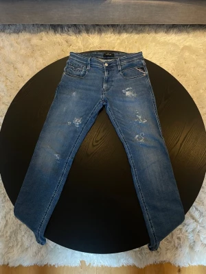 Blå slitna jeans Replay - Säljer ett par blå Replay Anbass jeans med slitningar och lätt tvättad look. Klassisk femficksmodell med raka ben och låg midja. Snygga detaljer på bakfickorna och Replay-logga fram. Perfekta för en avslappnad och trendig stil. 