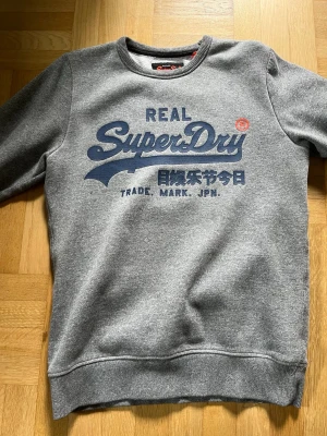 Grå sweatshirt från Superdry - Säljer en grå sweatshirt från Superdry med blå logga och japansk text på bröstet. Tröjan har rund halsringning, ribbade muddar och är tillverkad i mjukt material. Perfekt för en avslappnad och stilren look. Bara provat den en gång 