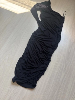 Svart asymmetrisk maxiklänning - Snygg svart maxiklänning med asymmetrisk axel och rynkade detaljer längs hela klänningen. Klänningen har en smal axelrem på ena sidan och ärmlös design på andra. Perfekt för dig som vill sticka ut med en unik look.