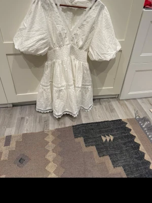 By malina klänning - Söt vit kortklänning med puffärmar, v-ringning och smock i midjan. Klänningen har spetsdetaljer längs kanterna och ett diskret prickigt mönster i tyget. Perfekt för sommarens alla tillfällen. Använd endast på studenten. 