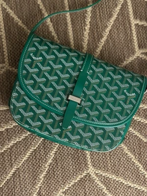 Grön Goyard handväska med mönster - Snygg grön handväska från Goyard med det klassiska vita och beigea Goyard-mönstret. Väskan har ett justerbart axelband och stängs med ett dekorativt spänne i silverfärg. Tillverkad i canvas med läderdetaljer. Perfekt för dig som vill sticka ut med en ikonisk accessoar.