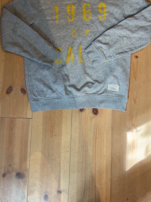 Grå sweatshirt från gap - Säljer en grå sweatshirt från gap med trycket '1969 S-F CALIF.' i gul text på bröstet. Tröjan har långa ärmar, rund halsringning och ribbade muddar vid ärmslut och nederkant. Materialet är mjukt och bekvämt, perfekt för en avslappnad stil.