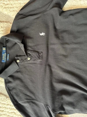 Svart långärmad piké från Ralph Lauren - Klassisk svart långärmad pikétröja från Ralph Lauren med vit broderad logga på bröstet. Tröjan har krage, två knappar vid halsen och är tillverkad i mjukt bomullstyg. Perfekt för en stilren och avslappnad look. Sitter som M men är L