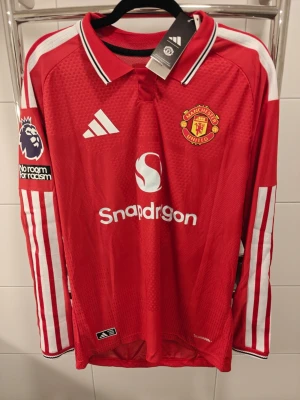 Player version nya Manchester United långärmad matchtröja - Röd långärmad Manchester United matchtröja från Adidas med vita detaljer och klubbmärke på bröstet. Tröjan har trycket 'B. Fernandes 8' på ryggen och Premier League-märke på ärmen. Tillverkad i lätt och ventilerande material.