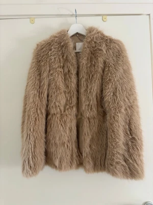 Beige fluffig pälsjacka - Säljer en beige, fluffig tunn  pälsjacka med lång lugg och rak passform. Jackan har en öppen front utan synliga knappar och är tillverkad i fuskpäls som ger en lyxig och trendig look. Perfekt för att hålla sig varm och snygg under kalla dagar. Kan knäppas eller ha öppen.