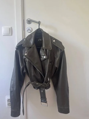 Skinnjacka med bälte från Zara - Snygg  skinnjacka från Zara med klassisk bikerstil, breda slag, silverfärgade detaljer och bälte i midjan. Jackan har dragkedja framtill.