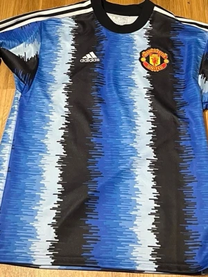 Manchester United blå Adidas tröja - Säljer en blå och svart Manchester United tröja från Adidas med grafiskt mönster och klubbmärke på bröstet. Tröjan har vita ränder på axlarna och rund hals. Perfekt för dig som älskar fotboll och vill sticka ut på planen.