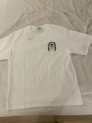 Vit Casablanca T shirt - Säljer en vit t-shirt från Casablanca med ett färgglatt tryck på bröstet och klassisk rund hals. T-shirten har korta ärmar och är gjord i mjuk bomull, perfekt för en avslappnad och stilren look.