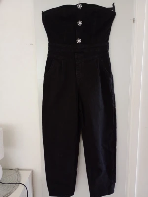 Svart strapless jumpsuit med knappar - Säljer en svart strapless jumpsuit med raka ben och höga fickor. Framtill har den dekorativa blommiga knappar och en markerad midja. Dragkedja bak för enkel påklädning. Materialet ser ut att vara denim eller kraftig bomull.
