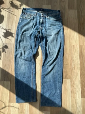 Blå jeans från GANT med raka ben - Säljer ett par klassiska blå jeans från GANT med raka ben och femficksdesign. Jeansen har en snygg ljusblå tvätt med subtila slitningar och GANT-logga på myntfickan. Materialet är denim i bomull och passformen är avslappnad. Storlek 34/30 men är justerade längst ner så mer 32/30 även lite lagade i skrevet men inget som syns/känns